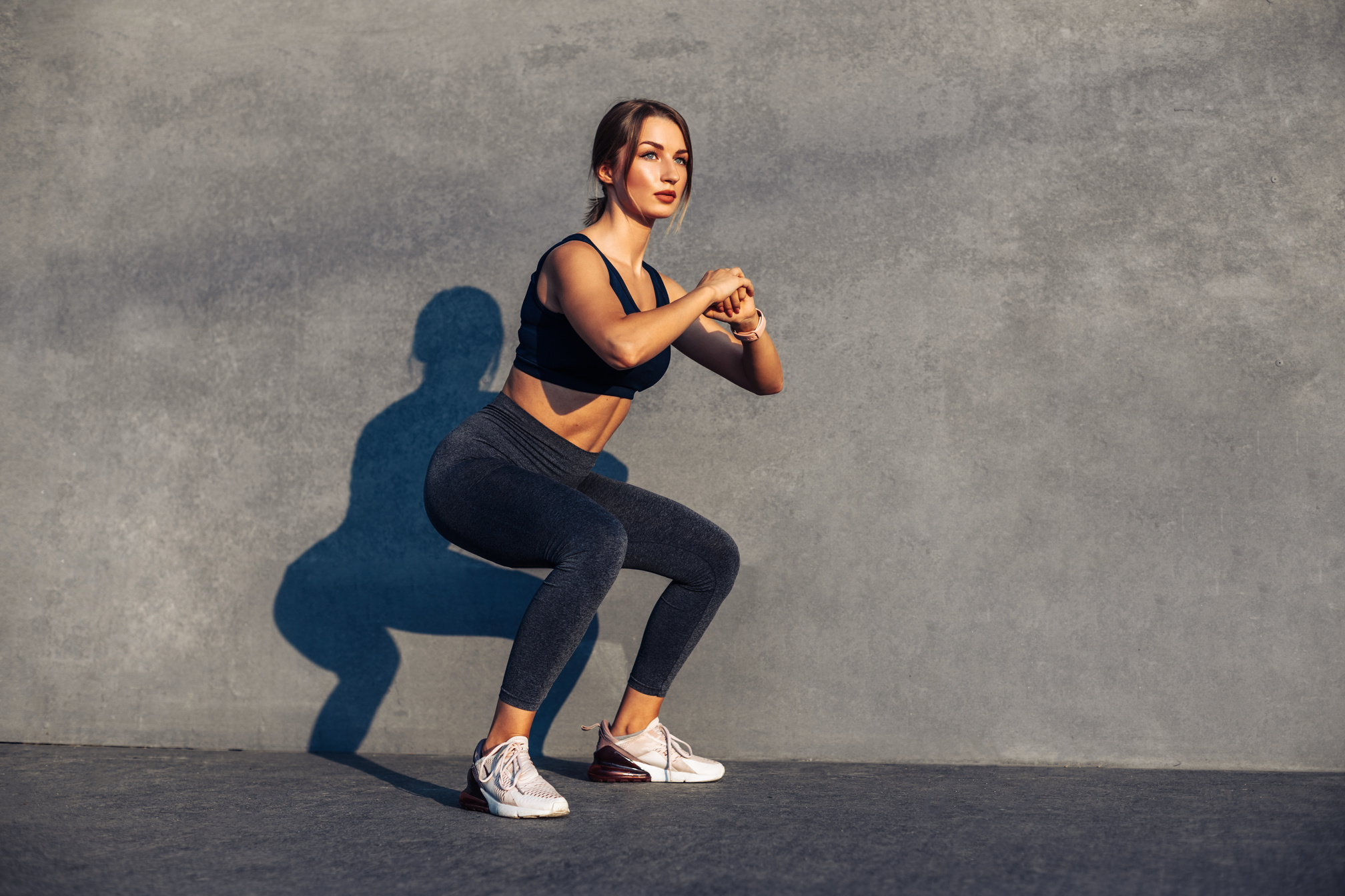 Sportive Woman, in Sneakers and a Tracksuit Squatting, Doing Squats on the Street Isolated against a Gray City Wall